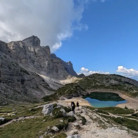 Two hikers descending a rocky trail toward a small turquoise alpine lake beneath grey limestone cliffs